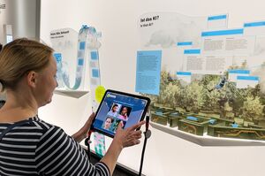 Eine Frau schaut auf ein Tablet. Im Hintergrund ist eine Infotafel zu KI zu sehen.