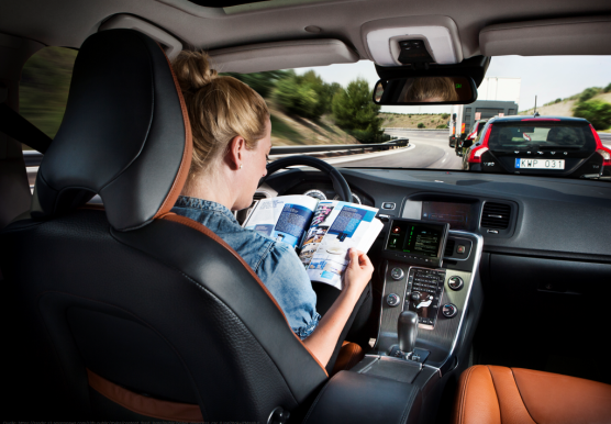 A woman sits at the wheel of an autonomously driving car. She is reading a magazine while driving.